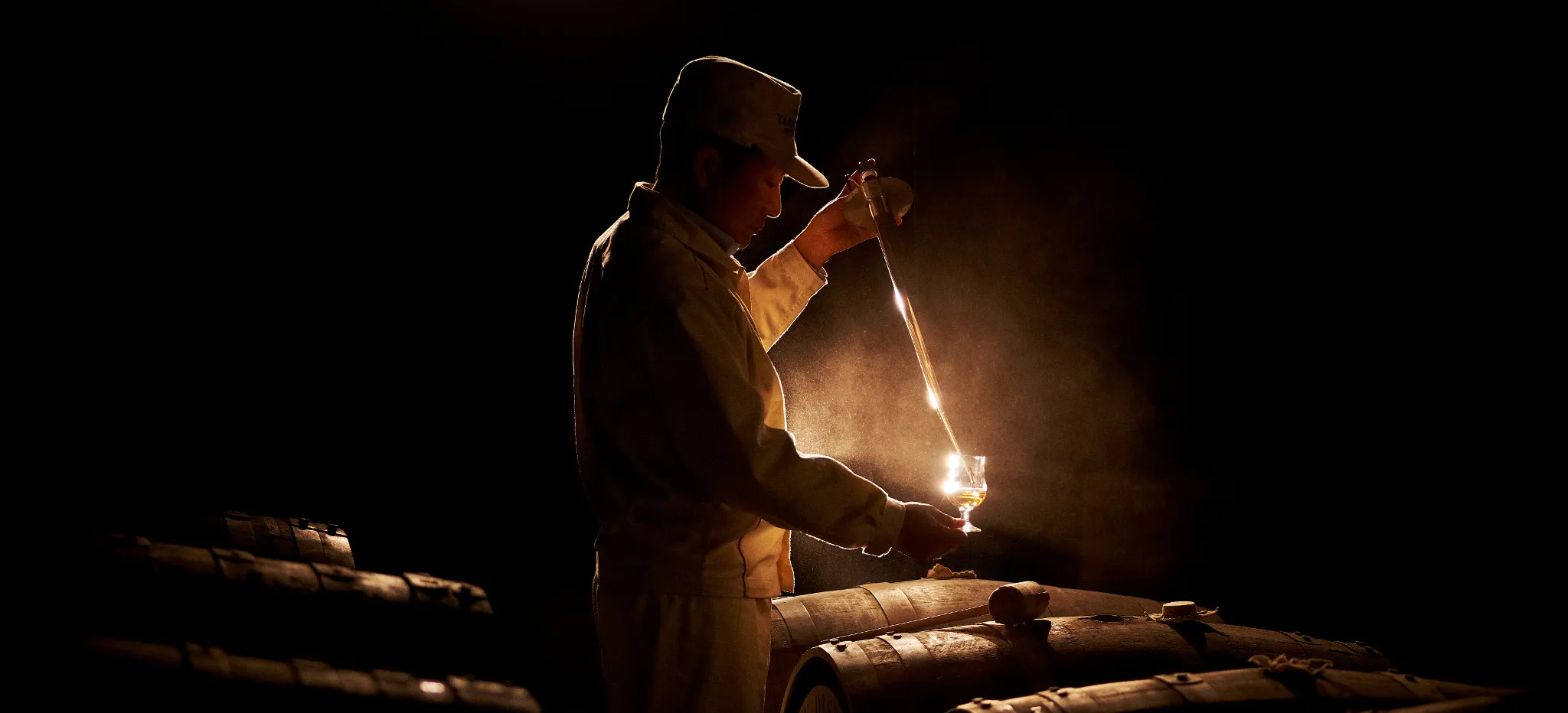 A Yamazaki distillery worker stands in a cellar surrounded by barrels. He expertly examines a glass of whisky. 